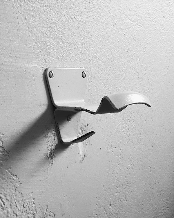 A double hook made of metal, gray paint, screwed to a wall. Found on a Sport Club in Apt, France, 2018.